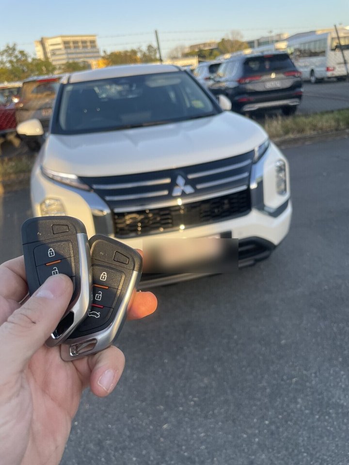Titan Automotive Locksmiths technician holding two freshly cut and programmed smart keys with the customer's Mitsubishi Outlander in the background at a Gold Coast dealership